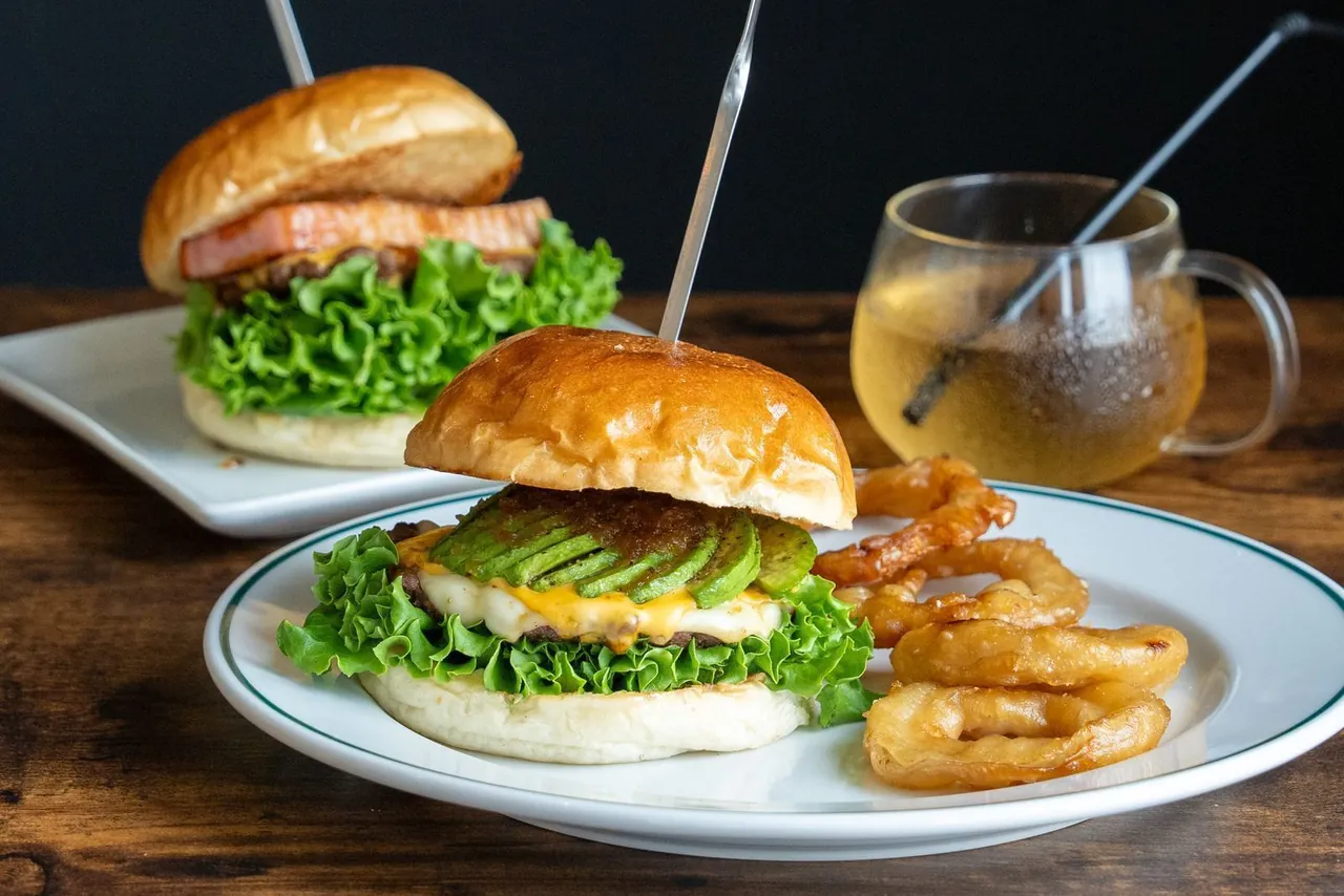 白い皿に2種のチーズとアボカドのバーガーとオニオンリング、奥に超厚切りベーコンチーズとドリンク