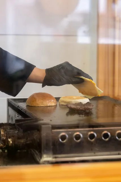 鉄板の上でこんがり焼けたバンズと、パティにチーズをのせる黒い手袋の手