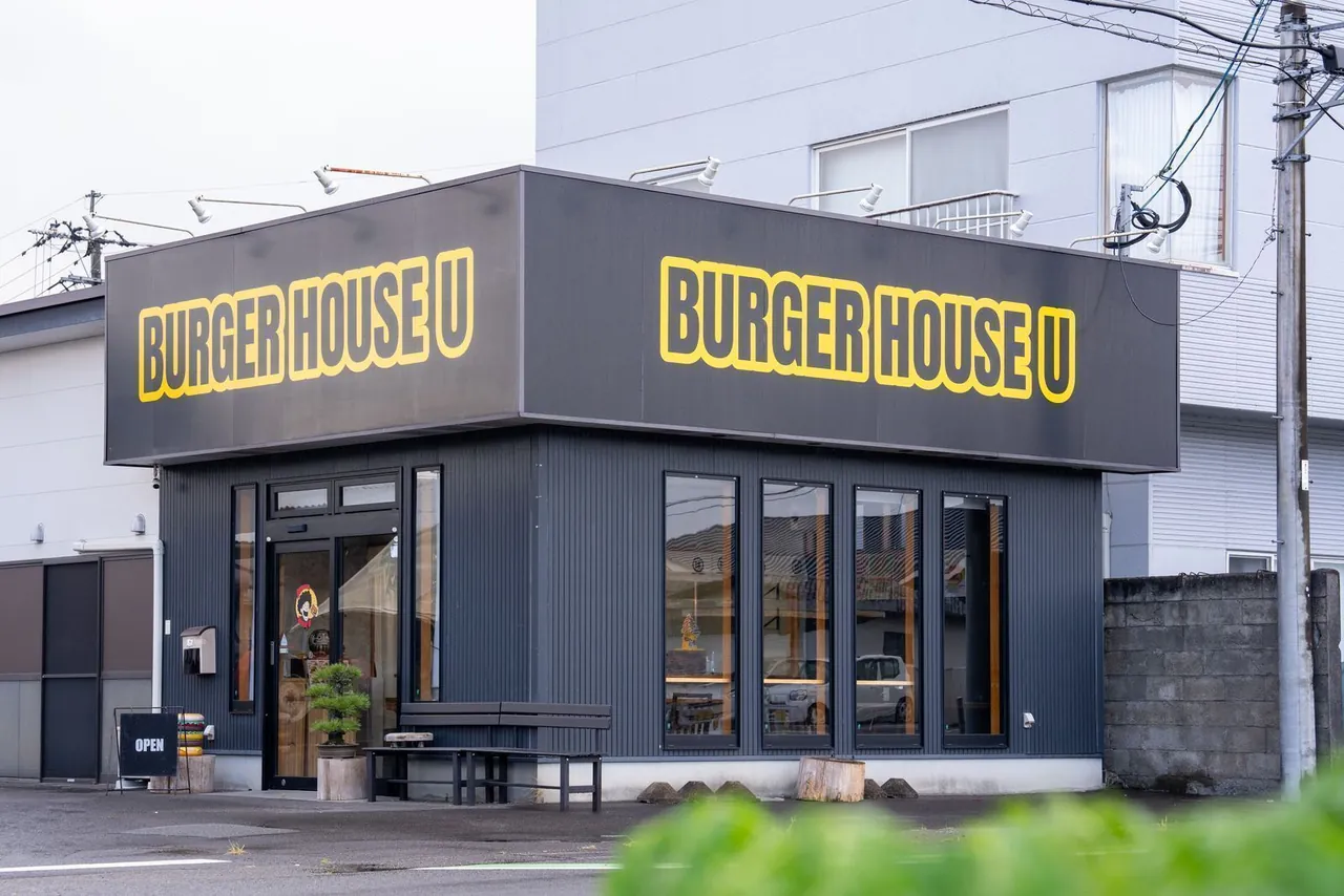 外観。黒い箱型の建物に、黄色い縁取りの「BURGER HOUSE U」の看板。入口に松の盆栽とベンチ、OPENの立て看板